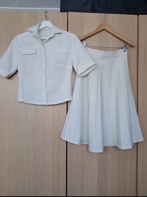 Vit kjol och skjorta set - Säljer ett stilrent set med vit kortärmad skjorta och matchande vid kjol. Skjortan har krage, knappar framtill och två bröstfickor med lock. Kjolen är midilång och har ett lätt utsvängd fall. Perfekt för en clean och fräsch look.