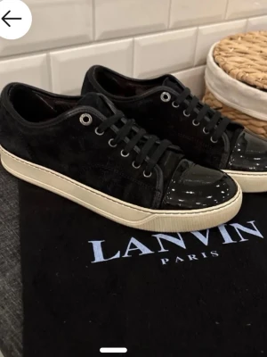 Svarta Lanvin sneakers med lackad tå - Snygga svarta sneakers från Lanvin med ovandel i mocka och en glansig, lackad tå. Skorna har svarta skosnören, silverfärgade öljetter och en vit sula som ger en clean kontrast. Perfekta för dig som gillar stilrena och exklusiva sneakers. liten defekt syns på sista bilden, enkel fix. 