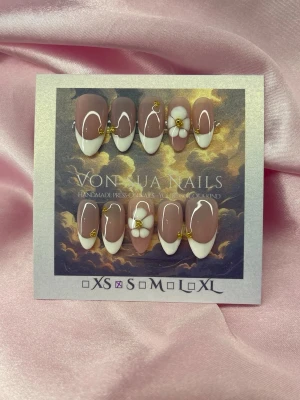 SoftBloom (S);Medium Almond - Snygga handgjorda press-on naglar i en medium längd och mandelform. Naglarna har en nude bas med vit fransk tipp, vita-blommor och små guldfärgade detaljer. Perfekt för dig som vill ha en elegant och trendig look. Handgjorda och unika i storlek S: 15 mm, 11,5 mm, 12 mm, 11 mm, 9 mm 