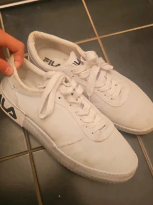 Vita Fila sneakers med logga - Säljer ett par klassiska vita sneakers från Fila med svart logga på sidan och plös. Skorna har lågt skaft, vita snören och en stilren design. Ovandelen är i textil och syntet, perfekt för dig som gillar en clean look.