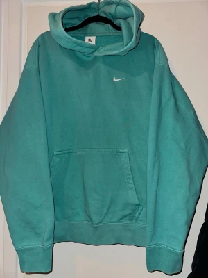 Turkos hoodie från Nike - Säljer en turkos hoodie från Nike med klassisk vit logga broderad på bröstet. Hoodien har en stor känguruficka framtill och en rejäl huva. Tillverkad i mjukt bomullsmaterial som är skönt mot huden. Perfekt för dig som gillar sportig och avslappnad stil.