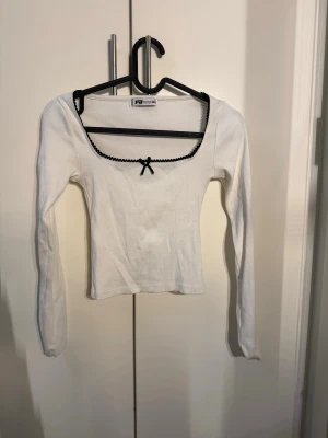 Vit ribbad topp med svart rosett - Söt vit ribbad topp från Fifth Avenue med lång ärm och fyrkantig ringning. Svart dekorativ kant och liten rosett framtill ger en gullig kontrast. Perfekt att styla med jeans eller kjol för en trendig look.