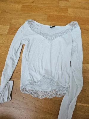 Vit långärmad topp med spets Gina Tricot - Säljer en vit långärmad topp från Gina Tricot med v-ringning och fina spetsdetaljer vid halsringning och nederkant. Toppen är ribbad och har en feminin, lite figurnära passform. Perfekt att styla med jeans eller kjol.