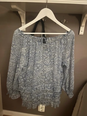 Blå mönstrad offshoulderblus - Blå offshoulderblus med vitt och blått mönster med långa ärmar. Blusen har resår vid axlar och ärmslut samt volangdetaljer nedtill. Tunn och luftig i materialet. 