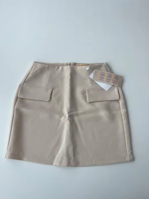 Beige kortkjol med fickor - Stilren beige kortkjol från Newyorker med två dekorativa fickor framtill och diskret dragkedja bak. Kjolen har en rak passform och är perfekt för en clean och modern look. Materialet känns mjukt och har en lätt struktur.