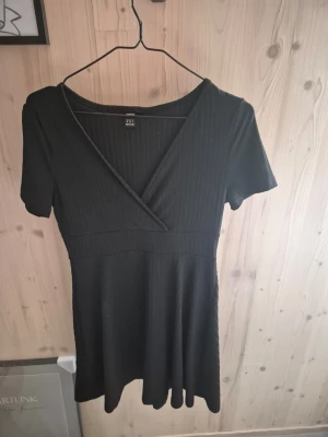 Svart omlottklänning  - En svart kortklänning med omlott framtill och v-ringad hals. Klänningen har korta ärmar och är tillverkad i ett ribbat material som ger en snygg struktur. Perfekt för dig som gillar enkel men stilren design.