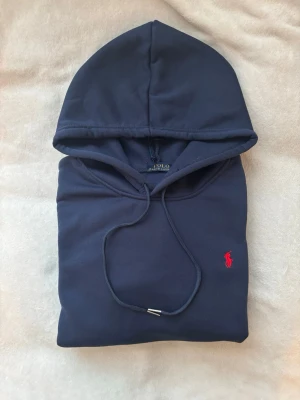 Mörkblå hoodie från Polo Ralph Lauren - Mörkblå hoodie från Polo Ralph Lauren med klassisk huva och dragsko. Tröjan har en känguruficka framtill och den ikoniska röda Polo-loggan broderad på bröstet. Tillverkad i mjuk bomullsblandning för skön komfort och stilren look.