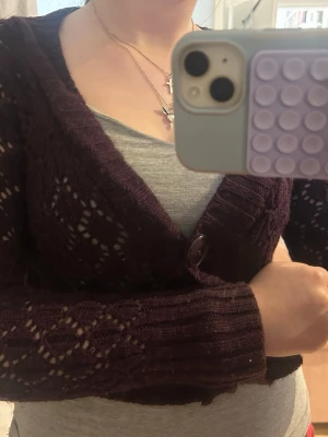  virkad kofta med stora knappar - Kort, plommonlila kofta med virkat mönster och två stora knappar framtill. Koftan har djup v-ringning och ribbstickade kanter. Perfekt för lager på lager och ger en trendig touch till din outfit.