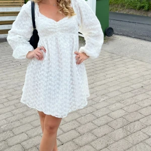 Vit kort klänning med blommönster - Söt vit kortklänning med subtilt blommönster och v-ringad hals. Klänningen har långa puffärmar och är tillverkad i ett lätt, luftigt material. Hade den som studentklänning så använd 1 gång enbart. Funkar som både storlek 38 och 36. Finns inte längre på NAKD 