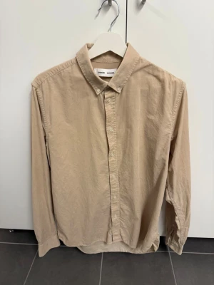 Beige skjorta från Samsoe Samsoe, stl L - Klassisk beige skjorta från Samsoe Samsoe i mjuk bomull. Skjortan har knappar framtill, button down-krage och långa ärmar med manschett. Perfekt basplagg med avslappnad passform och stilren look.