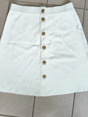 Vit kortkjol med knappar framtill ,Busnel - Snygg vit kortkjol med raka linjer och dekorativa guldfärgade knappar hela vägen framtill. Kjolen har en enkel och clean look som passar till det mesta. Tillverkad i ett mjukt bomullstyg för en bekväm känsla.