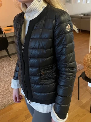 Svart dunjacka från Moncler - Snygg svart dunjacka från Moncler med quiltad design och rund krage. Jackan har dragkedja framtill, två fickor med dragkedja och Moncler-logga på ärmen. Perfekt för kalla vinterdagar och riktigt stilren look.