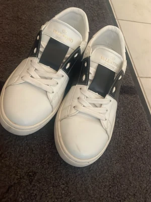 Valentino vita sneakers med svart detalj - Säljer ett par vita sneakers från Valentino med bred svart läderdetalj över snörningen och nitar på hälen. Skorna har klassisk låg profil och vita skosnören. Perfekta för dig som vill ha en stilren men edgy look.