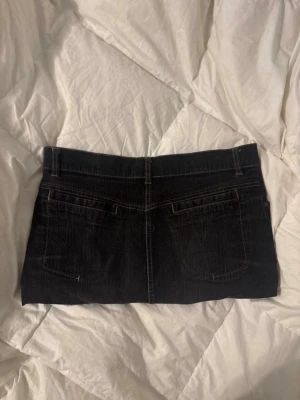 Svart jeanskjol i kort modell - Säljer en svart kort jeanskjol med klassiska fem fickor och bälteshällor. Kjolen har knapp och dragkedja framtill och är gjord i ett robust denimtyg. Perfekt för dig som gillar en enkel och stilren look.