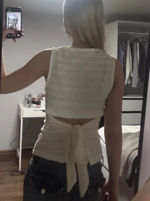 Vit ärmlös topp med knyt i ryggen - Säljer en vit ärmlös topp i strukturerat tyg med snygg knytning i ryggen. Toppen har en croppad passform och öppen rygg, vilket ger en trendig och luftig känsla. Perfekt till jeans eller kjol för en cool look.