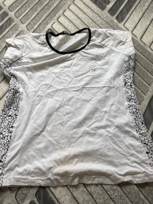 Vit t-shirt med svart mönster Redics - Vit t-shirt från Redics med svart kant runt halsen och svartvitt abstrakt mönster på sidorna. T-shirten har rund hals och är tillverkad i mjukt bomullsliknande material. Perfekt för en avslappnad och stilren look.its staind a bit