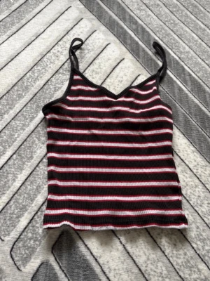 Randig ribbad linnetopp - Säljer en ribbad linnetopp med smala axelband och v-ringning. Toppen har svarta, vita och rosa ränder och är croppad i modellen. Perfekt för dig som gillar en enkel men snygg stil.