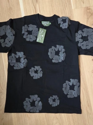 Svart t-shirt med grått blommönster från Denim Tears - Svart t-shirt från Denim Tears med gråa blommiga tryck över hela tröjan. Klassisk passform och rund halsringning. Tillverkad i mjuk bomull som känns skön mot huden. Perfekt för dig som vill sticka ut med en unik design.