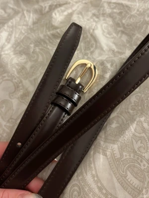 Mörkbrunt smalt skärp med gulddetalj - Snyggt och stilrent mörkbrunt skärp i skinn med en klassisk guldfärgad metallspänne. Perfekt till jeans eller för att piffa upp en outfit. Skärpet är smalt och har diskreta sömmar längs kanterna.