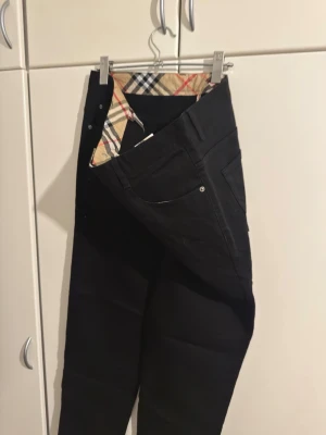Svarta  jeans med rutigt foder - Säljer ett par svarta jeans med det klassiska beige-rutiga fodret i midjan. Jeansen har fem fickor, raka ben och silverfärgade nitar. Materialet är kraftigt och har en exklusiv känsla. Perfekta för dig som gillar stilrena plagg med en twist. Har storlek w40 l34