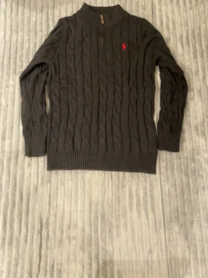 Polo Ralph Lauren Quarter Zip - Helt ny, taggar finns kvar. Kabelstickad svart tröja från Polo Ralph Lauren med halv dragkedja och ribbade muddar. Tröjan har en röd broderad logga på bröstet och hög krage. Perfekt för lager på lager och en klassisk look. Quarter Zip. Skickas samma dag.  