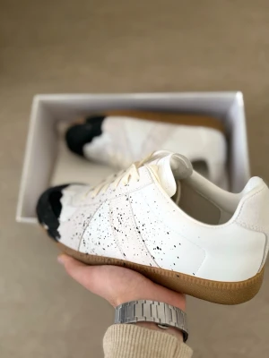 Maison Margiela sneakers vit/svart - Unika sneakers från Maison Margiela i vitt läder med svarta färgstänk och svart tåparti. Skorna har beige snörning och klassisk gummisula. 