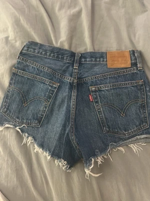 Levi's blå jeansshorts med fransar - Säljer ett par klassiska blå jeansshorts från Levi's med råa, fransiga kanter och femficksmodell. Shortsen har en vintagekänsla med medel midja och tydliga bakfickor med den ikoniska Levi's-lappen. Perfekta för sommaren och festivaler.