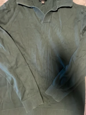 Grön stickad tröja från NEXT - Mörkgrön stickad tröja från NEXT med bred ribbad krage och v-ringning. Tröjan har långa ärmar och ribbade muddar vid ärmslut och nederkant. Perfekt för dig som gillar en enkel men snygg stil.