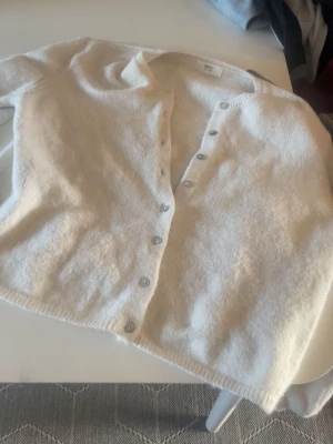 Vit kofta från Gina Tricot - Mysig vit kofta från Gina Tricot i alpackablandning. Koftan har långa ärmar, rund halsringning och knäppning framtill med silvriga knappar. Perfekt att slänga över en topp för en enkel och stilren look. Storlek XL men liten i storlek så mer M eller L