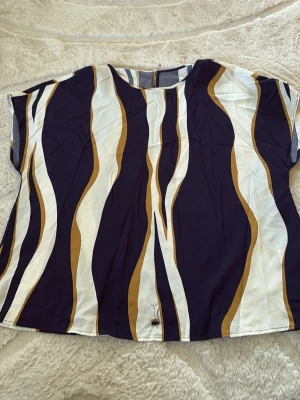 Randig topp i blått och gult - Snygg topp med vågiga ränder i mörkblått, vitt och senapsgult. Modellen är rak med korta ärmar och rund halsringning. Tillverkad i ett lätt och mjukt material som känns skönt mot huden. Perfekt för dig som gillar färg och mönster.