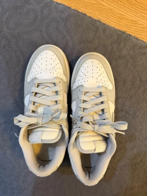 Nike Dunk Low vit/grå sneakers - Nike Dunk Low sneakers i vitt och ljusgrått läder med klassisk swoosh på sidan. Skorna har perforerad tåbox för ventilation, vadderad plös och krage samt matchande vita och grå skosnören. Perfekt för dig som gillar stilrena och tidlösa sneakers.