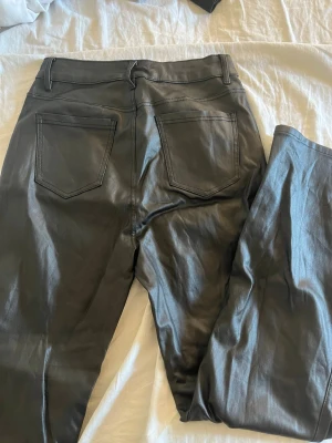 Svarta coated jeans med raka ben - Säljer ett par svarta coated jeans med raka ben och klassisk femficksmodell. Jeansen har hög midja och en snygg glansig finish som ger en cool look. Perfekta för dig som vill ha ett par trendiga byxor med skinnkänsla.
