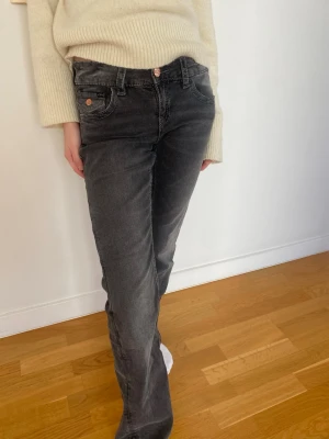 Grå raka jeans med rosettdetalj - Säljer ett par grå jeans med rak passform och låg midja. Jeansen har en snygg rosettdetalj med stenar på bakfickan och kopparfärgade knappar. Tillverkade i mjukt denimtyg som är skönt att bära hela dagen.