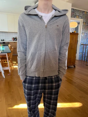 Grå zip hoodie från GANT 170 - Snygg grå hoodie från GANT med dragkedja och huva. Klassisk design med broderad logga på bröstet och två fickor framtill. Mjuk insida och ribbade muddar. Perfekt för en avslappnad och stilren look. Med ett litet hål vid fickan som knappast märks