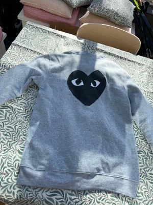 Grå hoodie Comme des cargons - M-L, Säljer en grå zip-hoodie från Comme des Garçons Play med ikoniskt hjärta med ögon på ryggen och en liten röd hjärtdetalj på bröstet. Hoodien har dragkedja, fickor fram och mjuk bomullskänsla. Perfekt för dig som gillar streetwear och snygga detaljer.