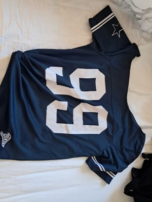 Blå Dallas Cowboys tröja - Säljer en blå självklippt Dallas Cowboys tröja med stora vita siffror 99 framtill. Tröjan har vita detaljer på ärmarna och en stjärna på axeln. Tillverkad i ett sportigt meshmaterial som andas, perfekt för träning eller match.