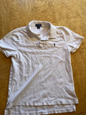 Vit pikétröja från Polo Ralph Lauren - Storleken är super off den passar 160-170. Klassisk vit pikétröja från Polo Ralph Lauren med korta ärmar och den ikoniska broderade loggan på bröstet. Tröjan har krage och knappar vid halsen. Perfekt till en avslappnad men ändå stilren look.