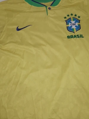 Brasilien fotbollströja Nike gul - Gul Brasilien fotbollströja från Nike med grönt och blått på kragen. Broderad CBF-logga och Nike Swoosh på bröstet. Klassisk design med knappar vid halsen och detaljer i grönt och blått. Perfekt för dig som älskar fotboll och vill stötta Brasilien.
