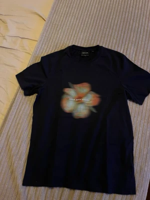 Blå t-shirt med blomtryck Lyle & Scott - Snygg mörkblå t-shirt från Lyle & Scott med stort blommigt tryck i orange och ljusblått på både fram- och baksidan. Klassisk passform och rund halsringning. Perfekt för dig som vill ha en unik och trendig look.