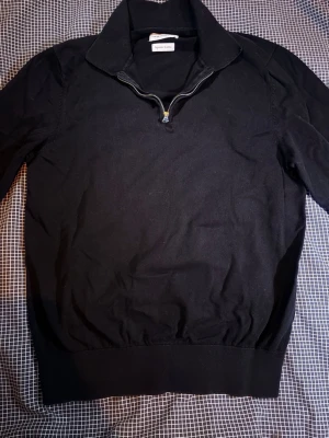 Svart halvzip tröja These Glory Days - Snygg svart tröja från These Glory Days med halvzip och hög krage. Tillverkad i mjuk bomull för skön känsla. Perfekt till jeans eller chinos för en clean look. Ribbstickad nederkant och ärmslut ger en schysst passform.