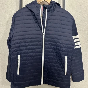 Thom Browné Jacket ~ 4 Bar - 🔹 Navy lightweight puffer jacket with a clean and sporty design  🔹 Quilted construction with horizontal stitching for a modern look and even insulation  🔹 Full front zip closure in white creating a sharp contrast against the navy fabric  🔹 Two zippered side pockets with white zippers for secure storage and visual balance  🔹 Comfortable hood that adds extra protection against wind and cooler weather  🔹 Sport-inspired double white stripe detail on the sleeve