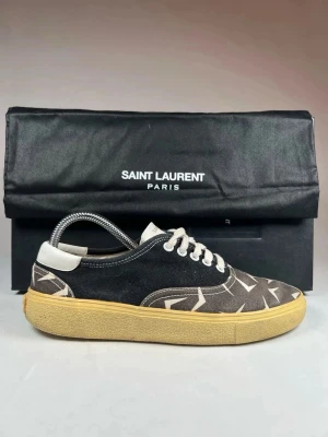 Saint Laurent svarta låga sneakers - Snygga låga sneakers från Saint Laurent i svart canvas med vita grafiska mönster. Skorna har vita snören, vit hälflik och en beige gummisula som ger en retrovibe. Perfekta för dig som gillar stilrena men ändå unika detaljer. Kommer med original box och dust bag, nypris 7900