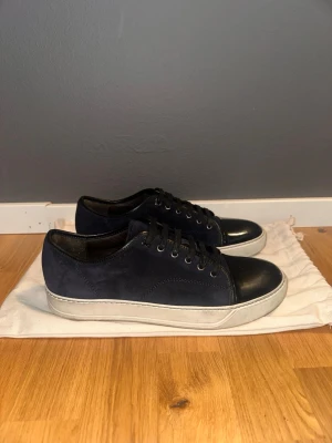 Mörkblå Lanvin  sneakers med svart tå - Snygga sneakers i mörkblå mocka med svart tåparti i läder och vita sulor. Skorna har svarta skosnören och metallfärgade öljetter. Perfekta för dig som gillar stilrena och klassiska sneakers med en twist.