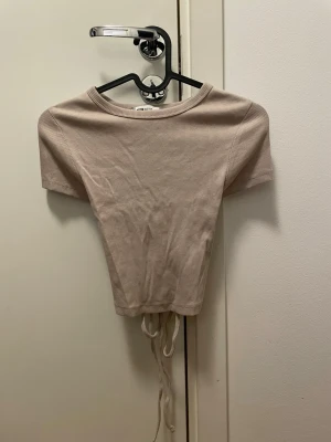 Beige ribbad croppad topp med knyt - Trendig beige croppad topp med korta ärmar och ribbad struktur. Toppen har knytband i nederkanten för justerbar passform och en enkel, stilren look. Perfekt att matcha med höga jeans eller kjol.
