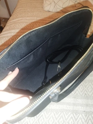 Svart datorväska med axelrem - Snygg svart datorväska med guldfärgad dragkedja och avtagbar axelrem. Väskan har ett stort huvudfack, innerficka och dekorativt mönstrat foder. Perfekt för laptop och tillbehör. Stilren design som passar till det mesta.