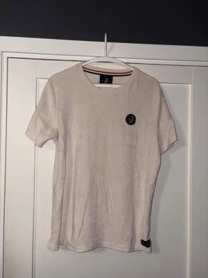 Beige frotté t-shirt från ACE - Säljer en beige t-shirt från ACE i mjuk frotté. T-shirten har korta ärmar och en rund halsringning. På bröstet sitter en patch med ett motiv av ett ansikte och nere vid fållen finns en liten ACE-logga. Perfekt för en chill och avslappnad stil.