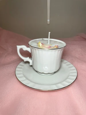 Doftljus i vanilj i en vit tekopp med fat  - Det här ljuset är gjort av återanvändbara material. Tekoppen är köpt secondhand och stearinet är från kyrkor som tänkte slänga det. Det här ljuset har en svag doft utav vanilj, så känsliga även kan använda det. Det här ljuset har även torkade blommor på som dekoration. 