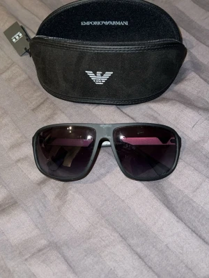 Svarta solglasögon Emporio Armani - Snygga svarta solglasögon från Emporio Armani med klassisk sportig design. Bågarna är mattsvarta och glasen är mörka, vilket ger en stilren look. Kommer med originalfodral med Emporio Armani-logga.