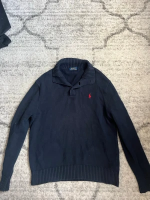 Mörkblå stickad tröja Polo Ralph Lauren - Mörkblå stickad tröja från Polo Ralph Lauren med ribbad krage och knappdetalj. Klassisk röd logga broderad på bröstet. Tröjan har långa ärmar och ribbade muddar vid ärmslut och nederkant. Perfekt för en stilren och avslappnad look.
