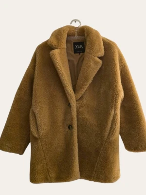 Brun teddyjacka från Zara - Säljer en supermjuk teddyjacka från Zara i en snygg brun färg. Jackan har bred krage, två knappar framtill och sidofickor. Oversized modell med fluffigt material som ger en riktigt cozy vibe. Perfekt för kalla dagar när du vill vara både varm och trendig.